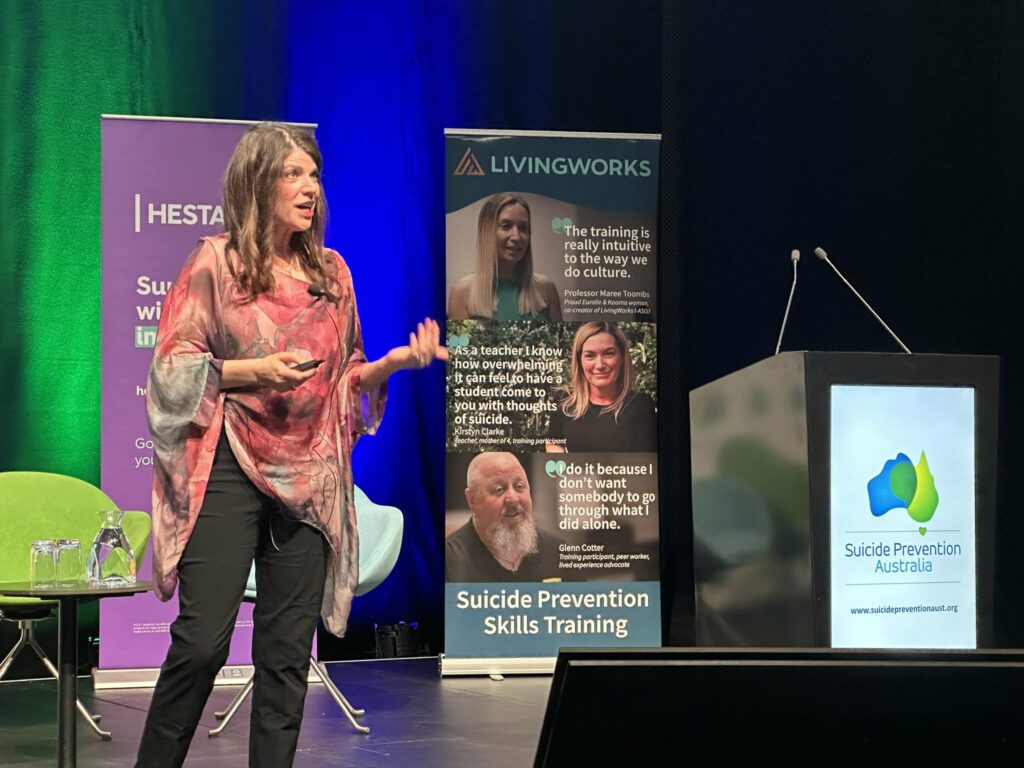 Dr Laura Shannonhouse standing on a stage at NSPC 2024 in Adelaide, behind her is a podium and a LivingWorks suicide prevention skills training banner.